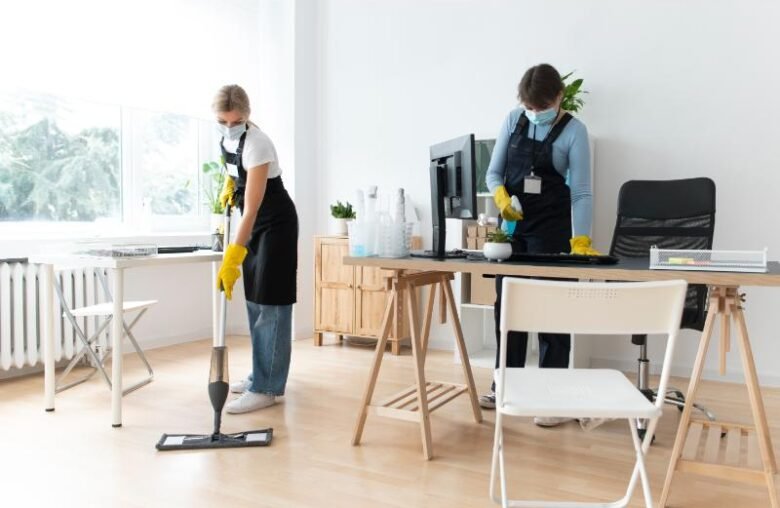 Two individuals in aprons cleaning an office space with a vacuum and cleaning supplies.