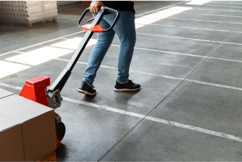 Person in blue jeans and black sneakers pulling a red hand pallet truck with cardboard boxes on a concrete floor.